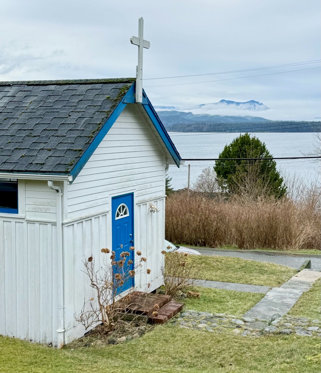 A white church with a blue door in front of the ocean with mountains in the background