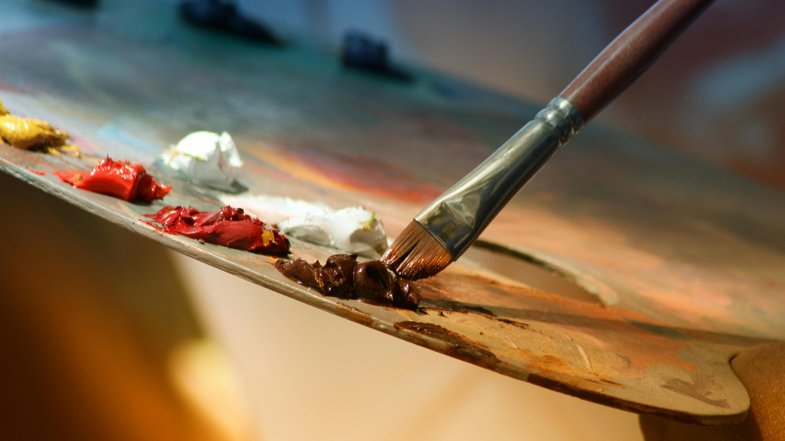 A close-up of an artist's wooden palette with various oil paint colors, including red, white, yellow, and brown. A paintbrush with a wooden handle and metal ferrule is dipped in dark brown paint, resting on the palette. The background is blurred, showing hints of a studio or creative workspace.