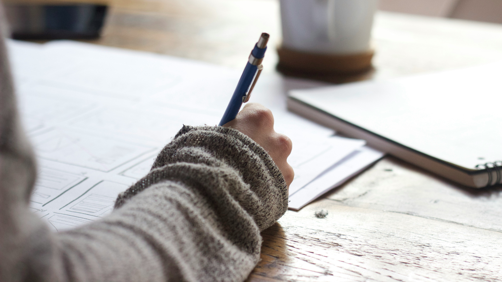 Person wearing a grey sweater writing with a pen on paper at a wooden desk, with a notebook and coffee cup nearby.