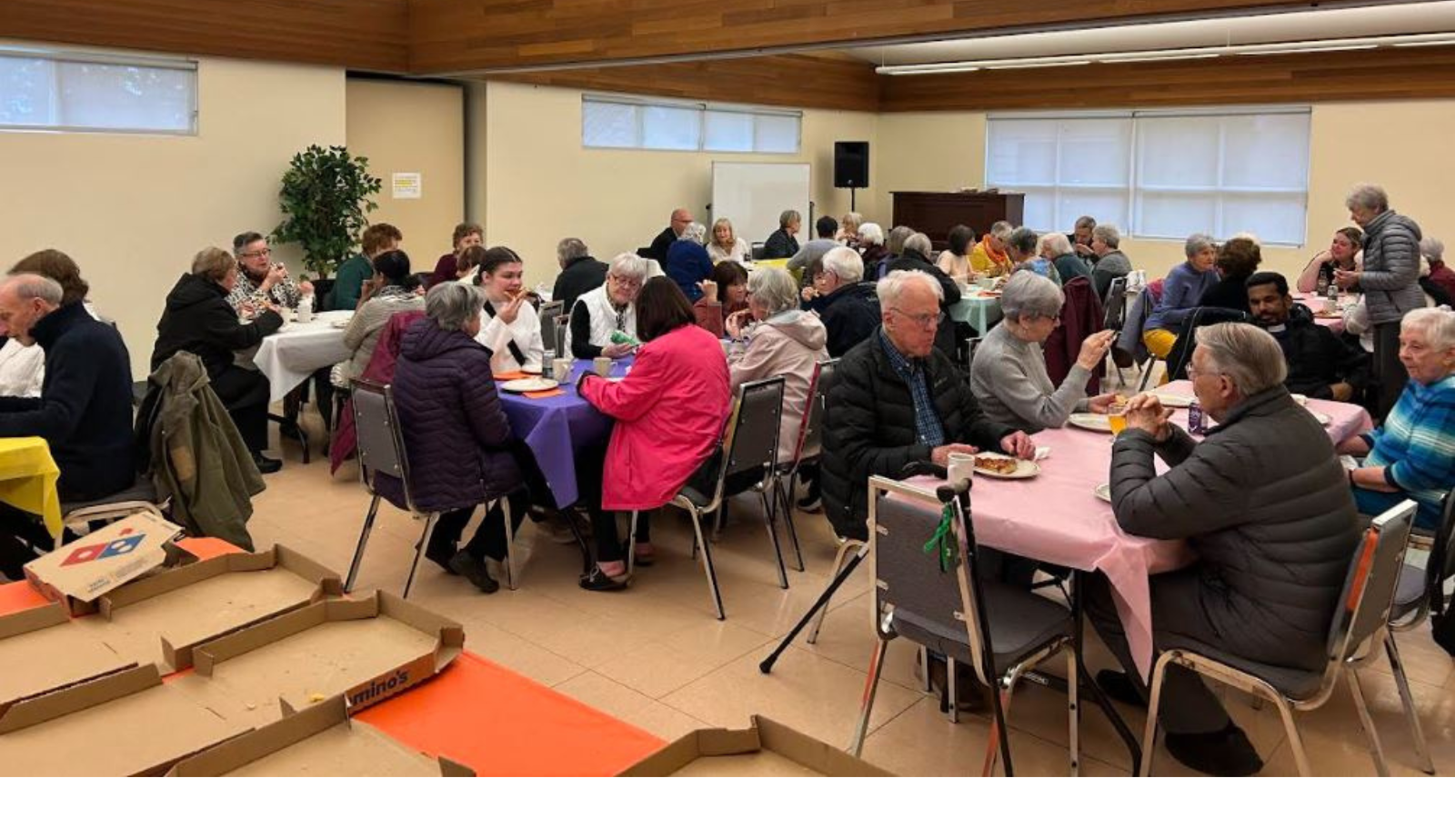 A lively gathering in a parish hall, where attendees enjoy pizza, conversation, and community. Empty Domino’s boxes sit in the foreground.