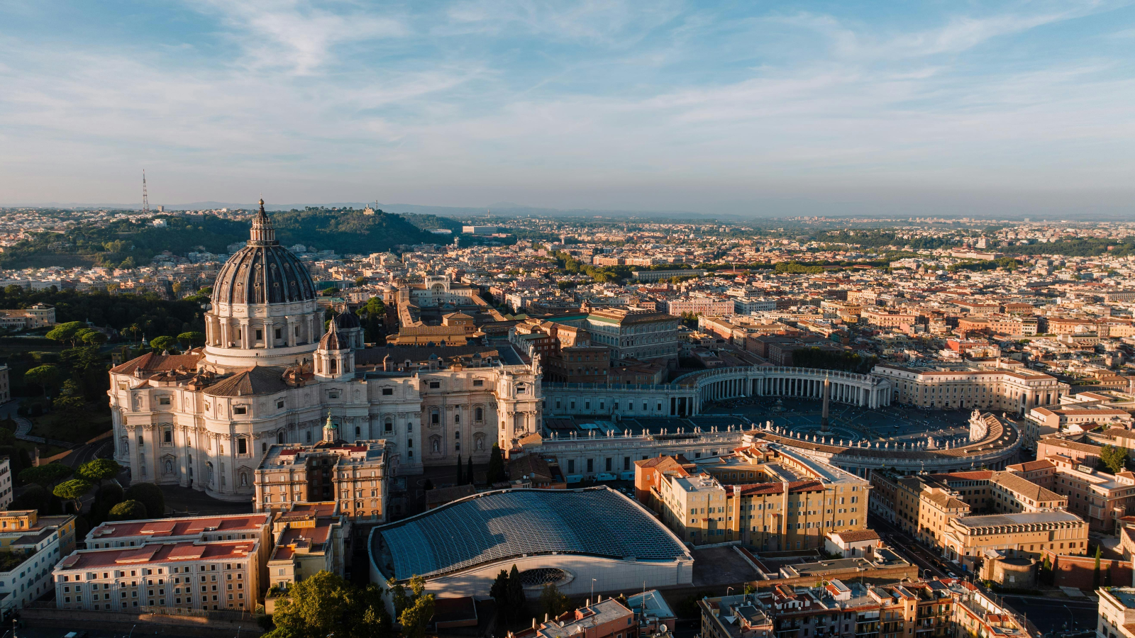 An aerial view of St. Peter's Basilica in Vatican City, featuring the grand dome and surrounding architectural details, with St. Peter's Square and its colonnades visible in the foreground. The urban landscape of Rome stretches into the horizon under a clear, sunny sky.