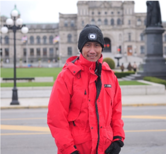 A man in a red jacket and black toque stands in front of the Victoria legislature buildings
