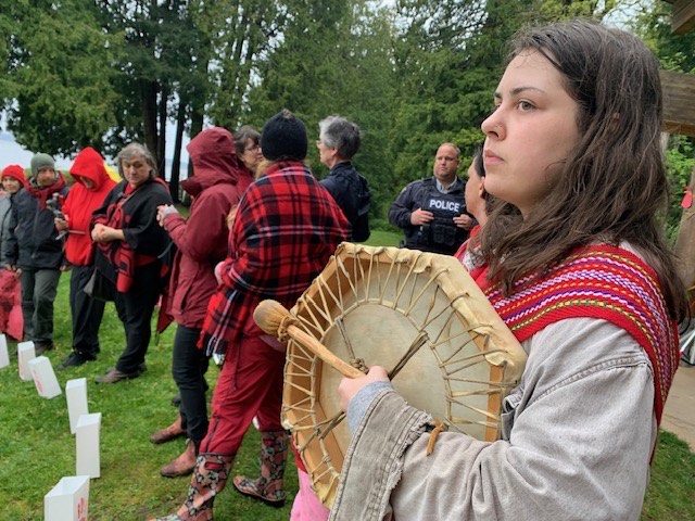 Photo of a girl standing outdoors, wearing a Metis sash and holding a drum, with other people in the background