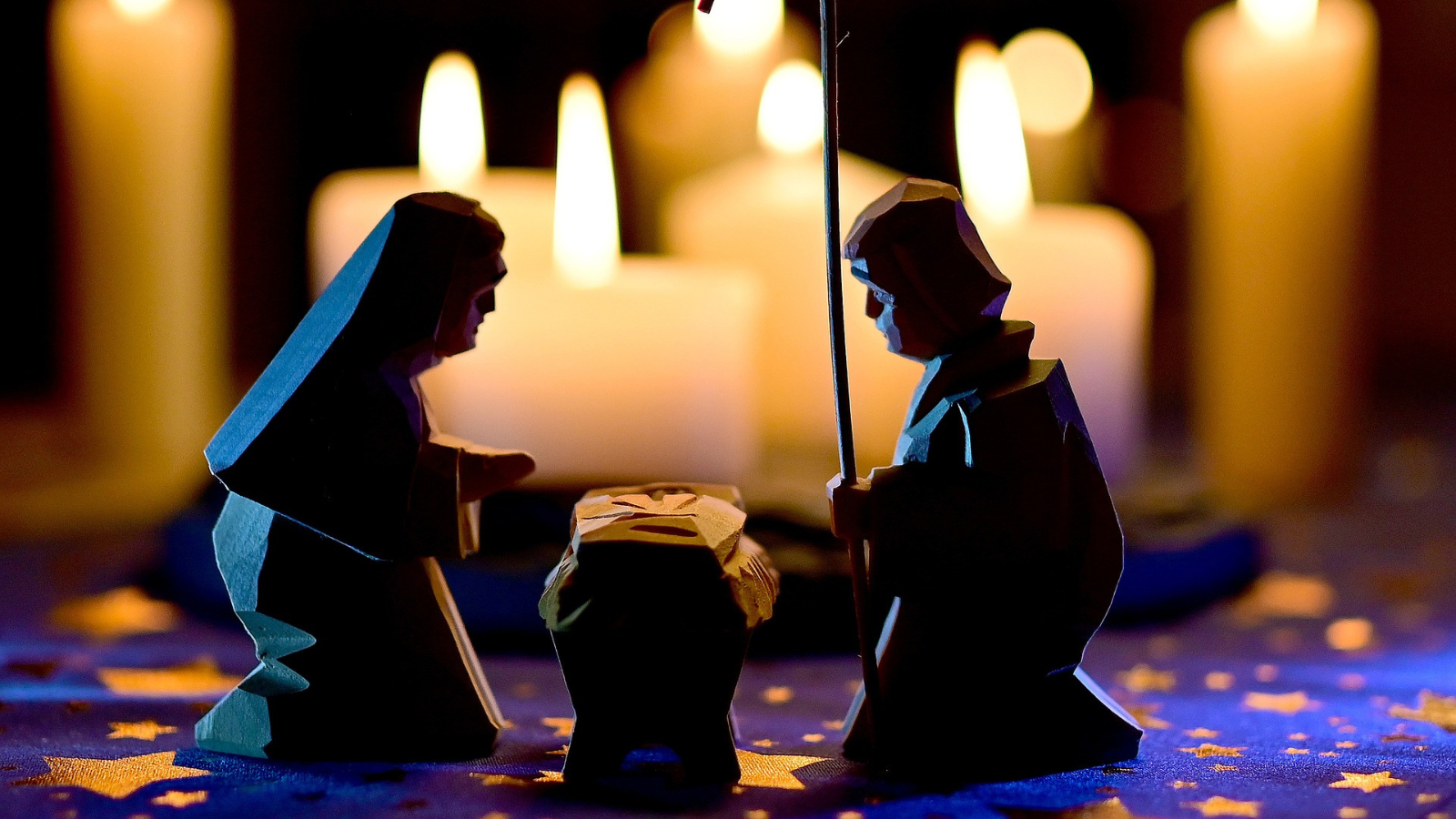 A close-up of a nativity scene with wooden figures of Mary, Joseph, and baby Jesus in a manger, softly illuminated by the warm glow of candles in the background. The foreground features a dark blue surface adorned with golden star patterns, creating a peaceful and festive atmosphere.