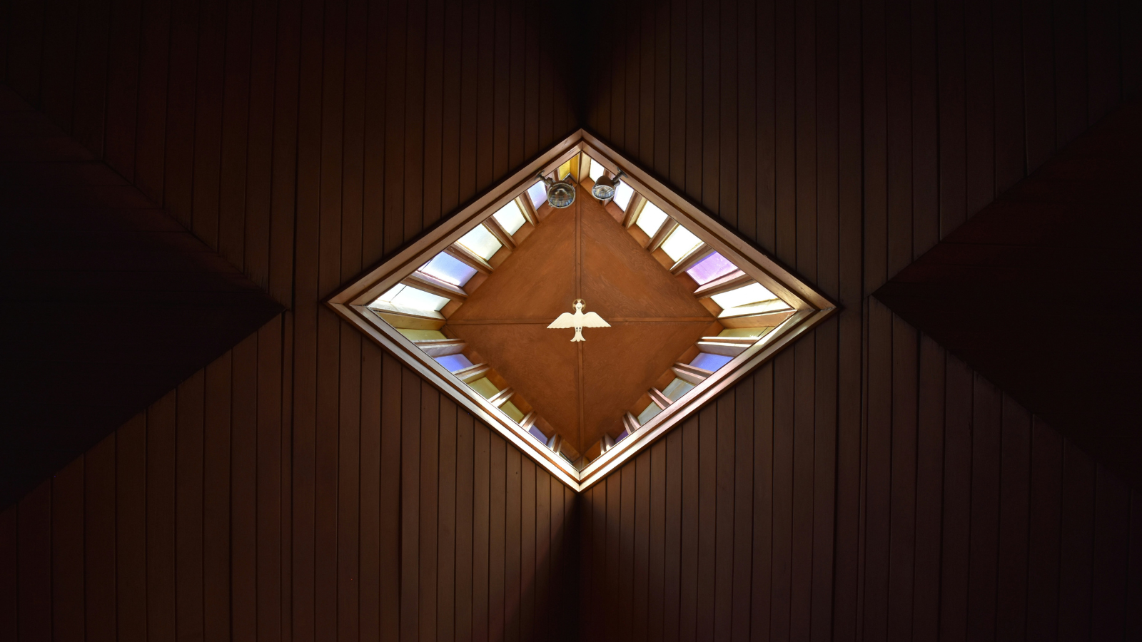 A view of a church ceiling featuring a diamond-shaped skylight with colorful stained glass panels. In the center hangs a white dove symbolizing the Holy Spirit, illuminated by soft natural light. The surrounding ceiling is made of dark wooden panels, creating a warm and reverent atmosphere.