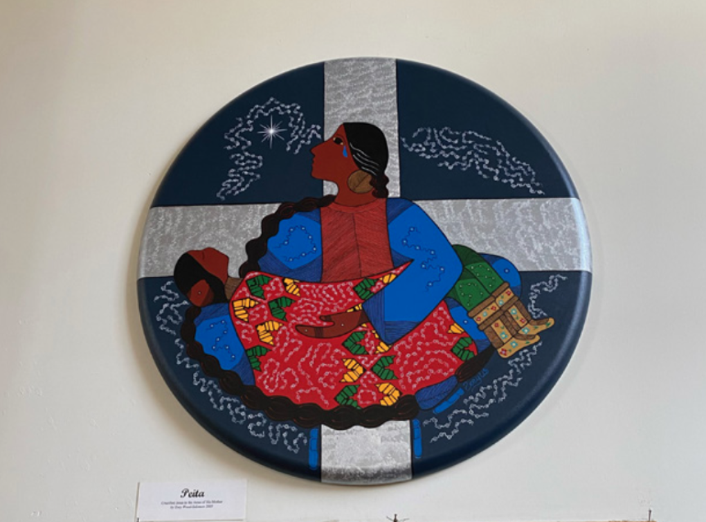 A photo of a circular painting of a crying woman holding a reclining man against the background of a cross, done in an Indigenous art style