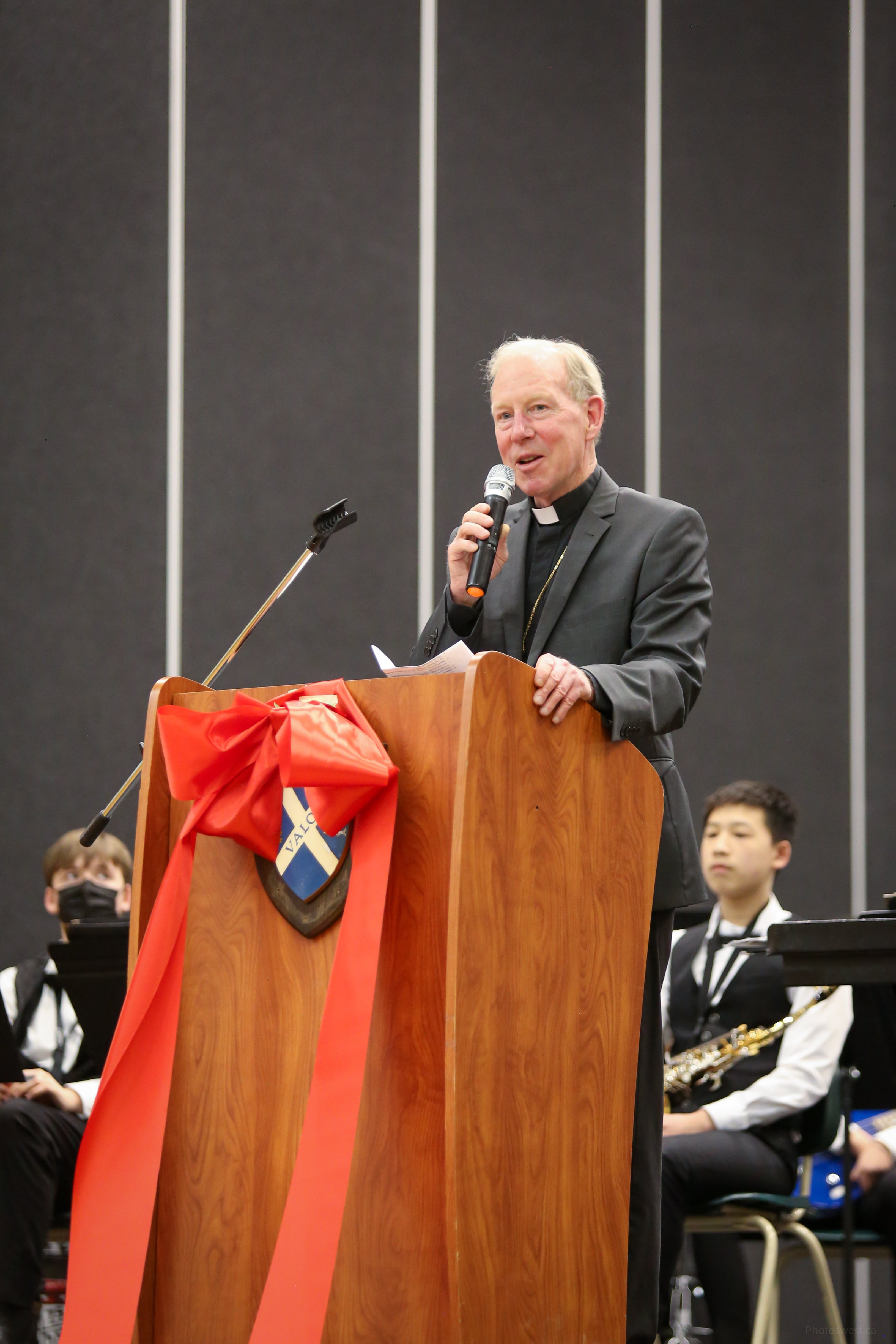 Bishop Gary speaking at a podium decorated with St. Andrew's Regional High School crest and a red bow, while students look on