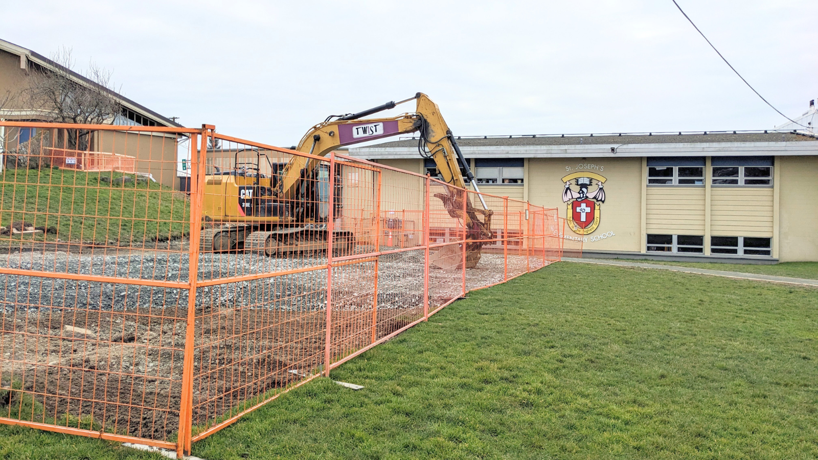 A construction site at St. Joseph’s Elementary School, enclosed by orange safety fencing. A large excavator is at work, preparing the ground for the new early learning centre. The school building with its emblem is visible in the background.
