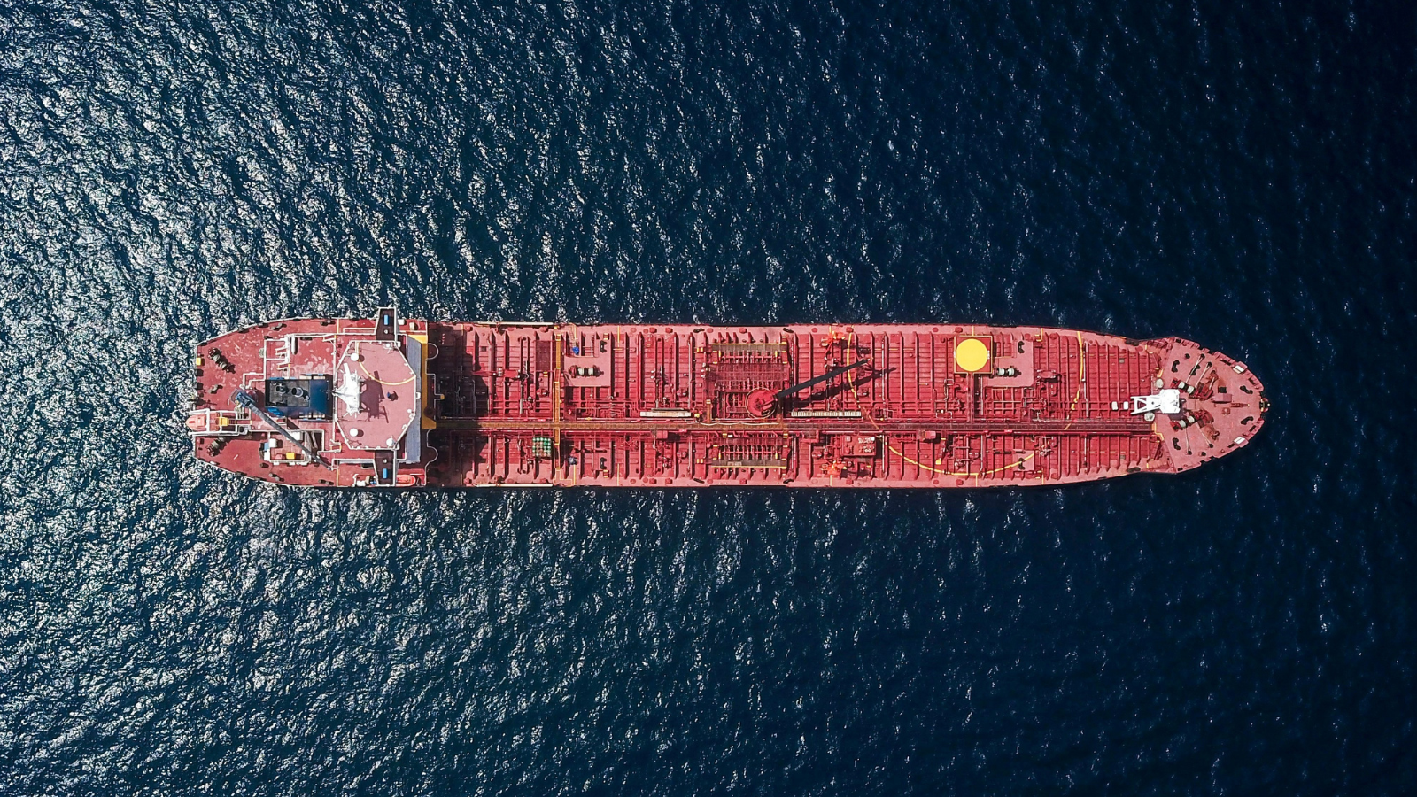 Aerial view of a large red cargo ship or oil tanker at sea, centered in deep blue ocean waters, with visible deck structures and equipment.