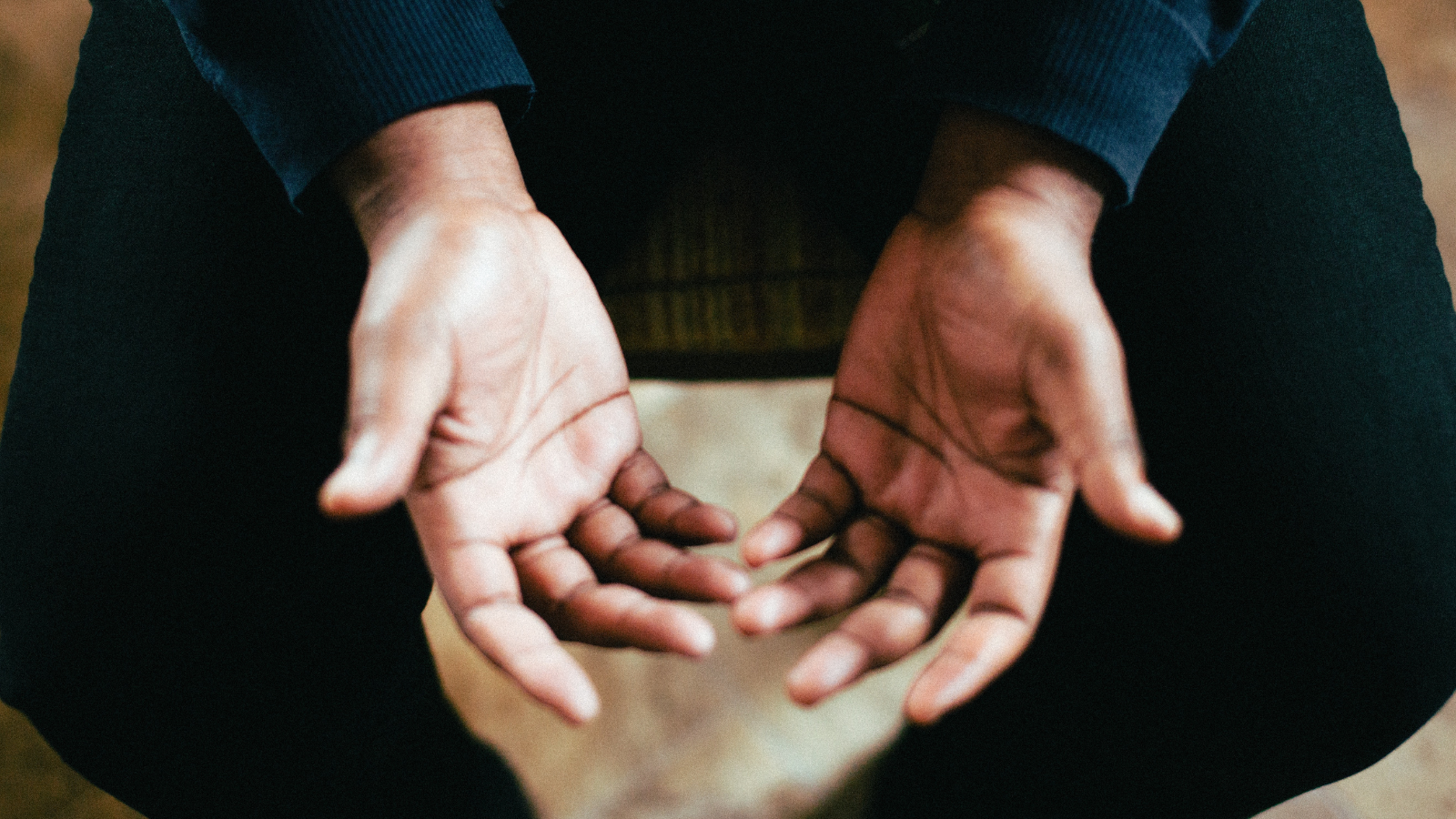 Close-up of a person’s open hands resting on their lap, palms facing up in a gesture of prayer, wearing a dark long-sleeve shirt and pants.