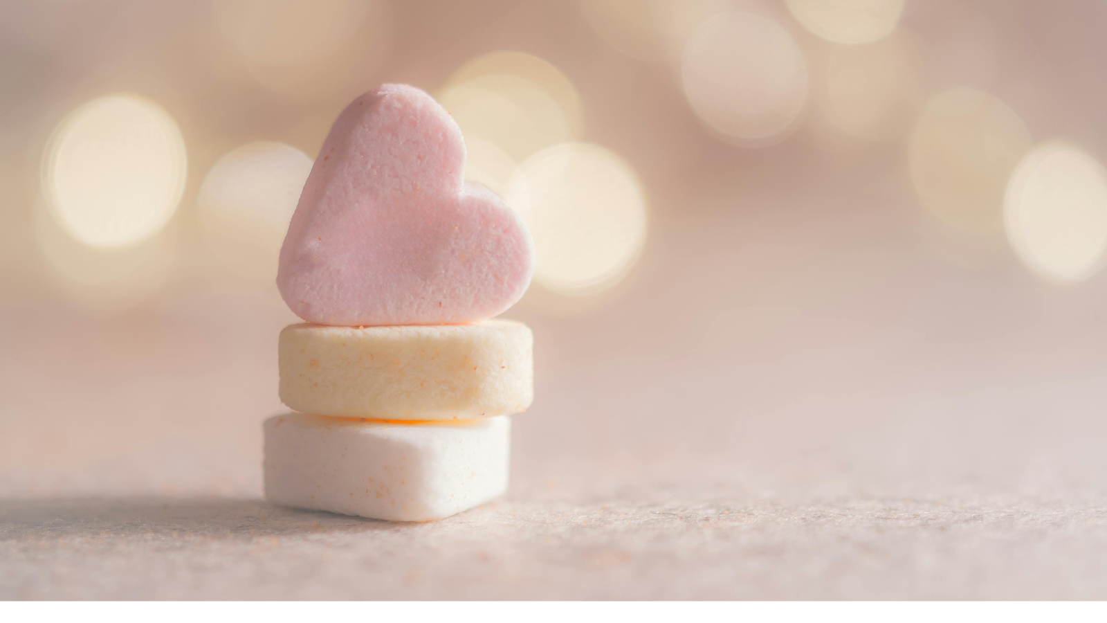 A stack of three pastel-colored candy hearts, with a pink heart-shaped candy on top of two round white and yellow candies. The background is soft and blurred with warm, glowing bokeh lights, creating a romantic and dreamy atmosphere.