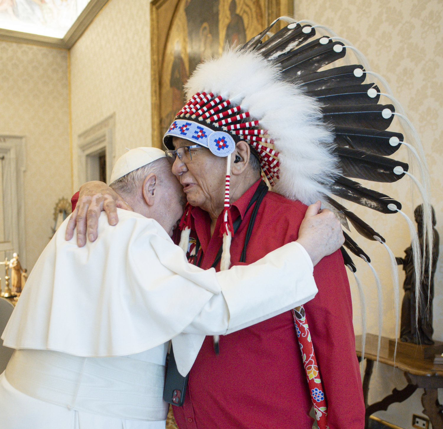 Pope Francis embracing an elderly Indigenous man who's wearing a feathered headdress