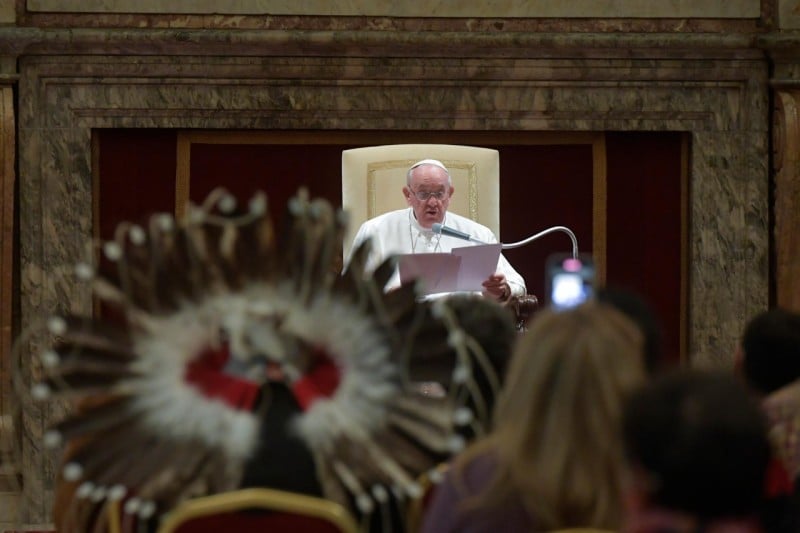 Pope Francis reading an address to an audience which includes a person in an Indigenous feathered headdress